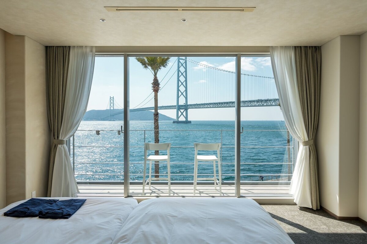 Ein Hotelzimmer mit großem Fenster, einem Bett, zwei Sesseln und Blick auf eine Brücke über dem Wasser.
