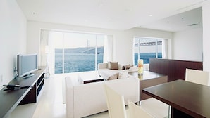 A modern living room with a large window overlooking a body of water and a bridge, a flat-screen TV on a stand, a sofa, and a dining table with chairs. - HOTEL SETRE KOBE MAIKO (Kobe)