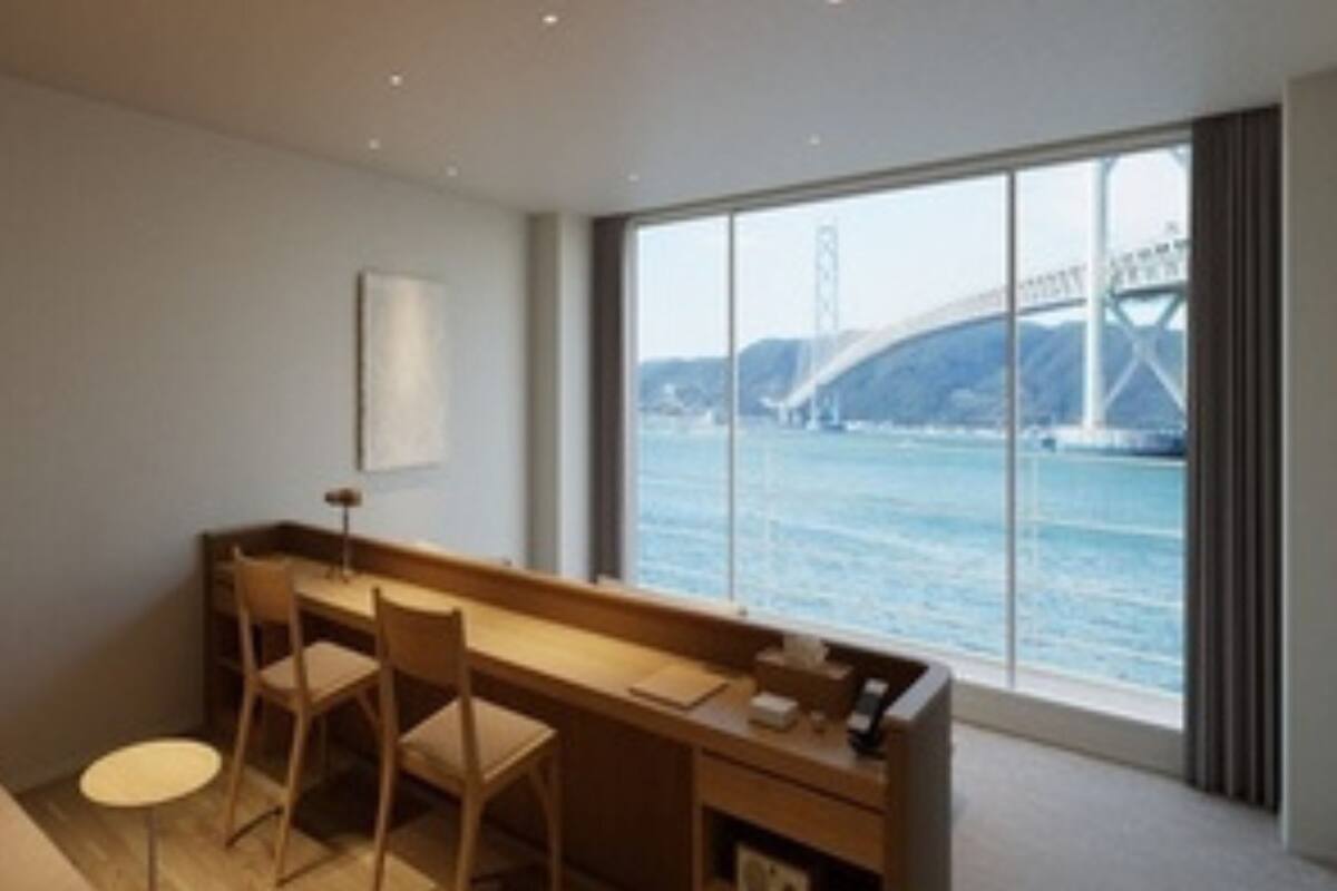 Une chambre d’hôtel moderne dotée d’une grande fenêtre donnant sur un pont et sur l’eau. On y trouve un bureau en bois avec deux chaises, une petite table ronde et un canapé.