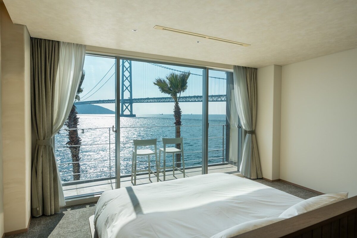 Ein Hotelzimmer mit großem Fenster und Blick auf eine Brücke sowie Wasser. Ein Bett, zwei Stühle und eine Palme sind von außen sichtbar.