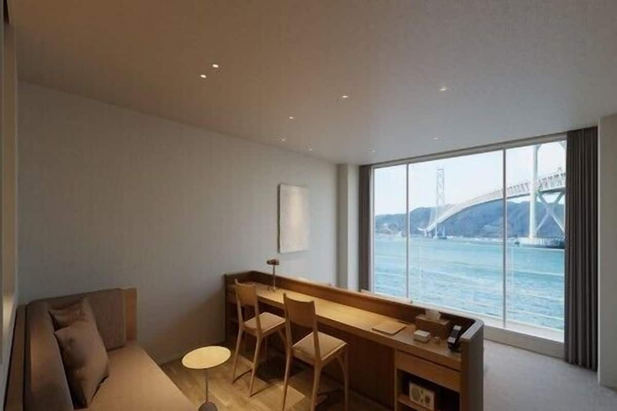 A modern hotel room with a sofa, a desk, and a view of a bridge over water.