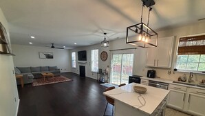 A modern kitchen with a central island, a dining area with a table and chairs, a living room with a sofa and a fireplace, and a large window with blinds.