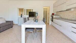 A modern kitchen with white cabinets and a built-in oven. There is a dining area with a white table and chairs. A gray sofa is visible in the background. - Villa Anna by Interhome (Castiglione della Pescaia)