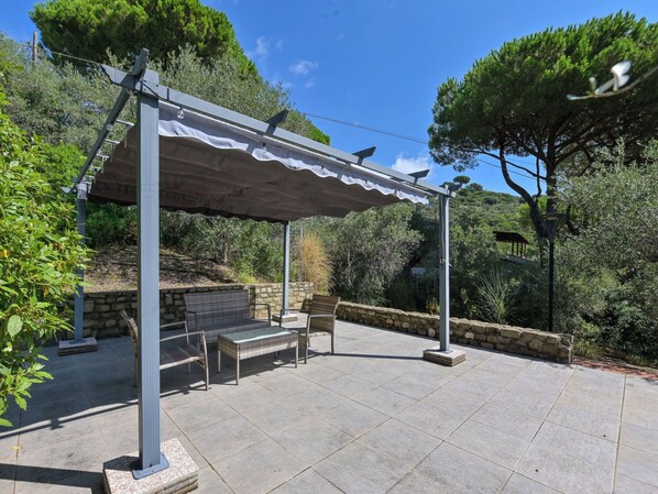 Une terrasse extérieure avec un sol en pierre, une pergola surmontée d’une auvent blanche, des meubles en osier et un mur en pierre.