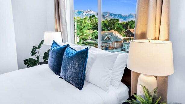 A hotel room with a bed, a lamp, and a view of mountains and a village. - Sugarhouse! Mountain View 3BR, 2BA Gym, Parking (Salt Lake City)