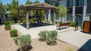 Un espace extérieur aménagé, abrité et doté de bancs et de tables de pique-nique, entouré d’arbustes décoratifs et bordé d’un bâtiment aux grandes fenêtres.