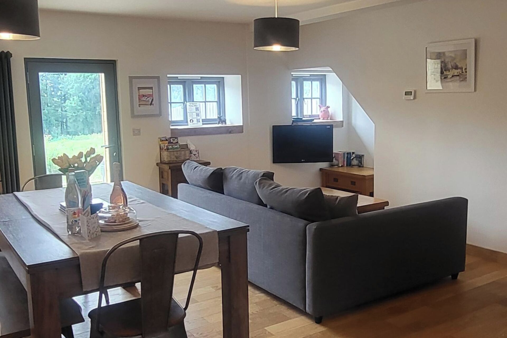 En moderne stue med en grå sofa, et spisebord med stole, et fladskærms-tv på væggen og et vindue med udsigt til et hus.