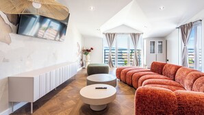 Ein modernes Wohnzimmer mit einer Couch, Poufs, einem Fernseher und einem großen Fenster mit Vorhängen.
