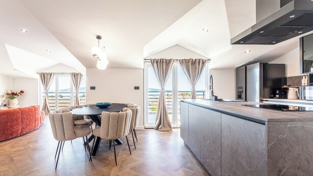 Eine moderne Küche mit einer großen Kochinsel, einem Essbereich mit rundem Tisch und Stühlen sowie Ausblick ins Freie durch große Fenster.