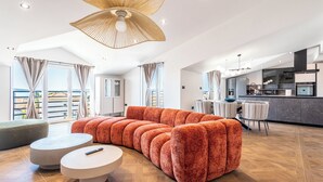 Ein modernes Wohnzimmer mit einem großen, orangefarbenen Sofa, einem runden Couchtisch und einem Essbereich, dessen Hintergrund eine Küche zeigt.