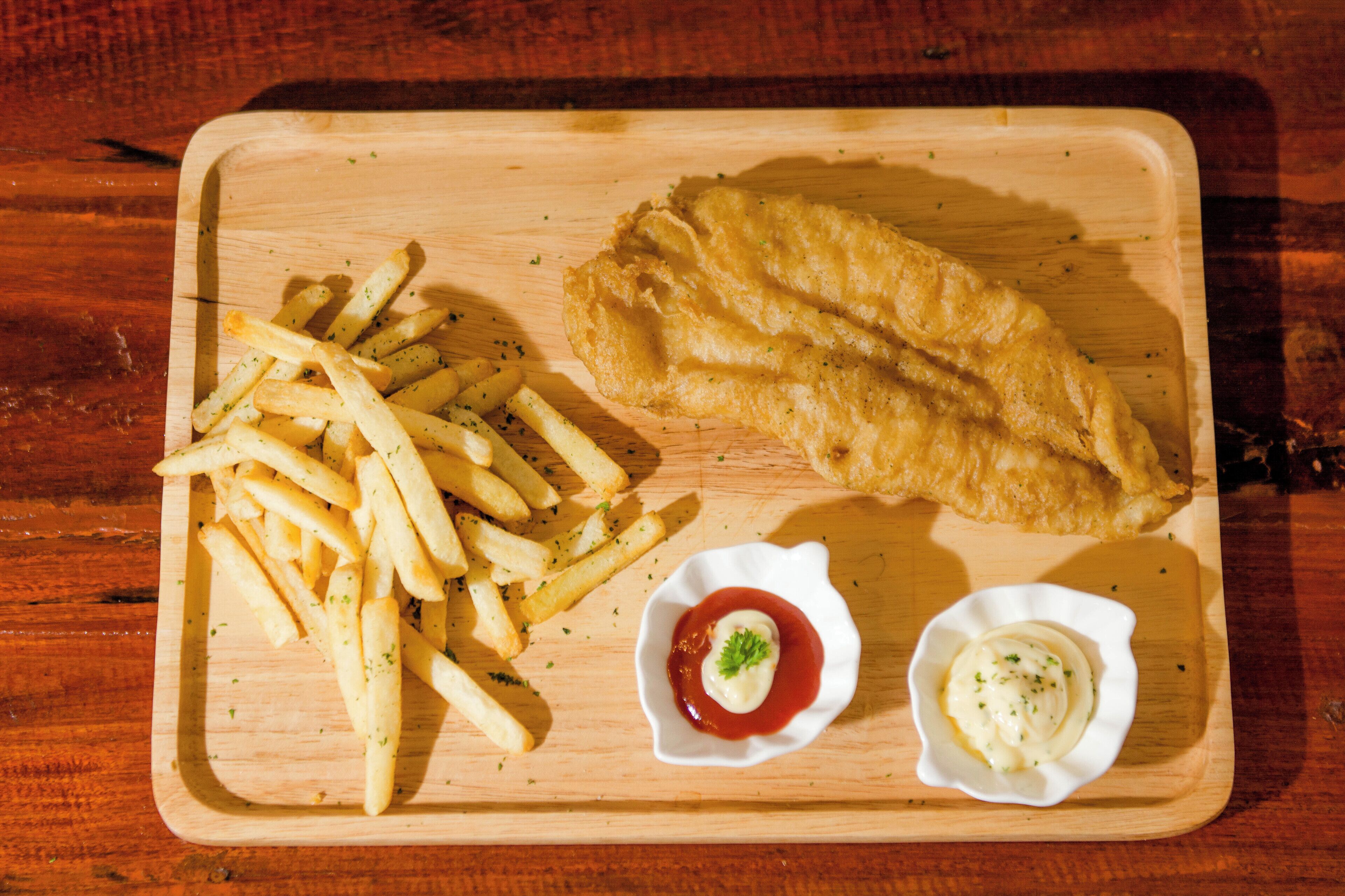 Un vassoio di legno con un filetto di pesce, patatine fritte e due piccole ciotole di salsa.