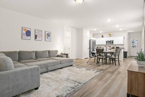 A modern living room with a grey sofa, a dining area with a table and chairs, and a kitchen in the background.