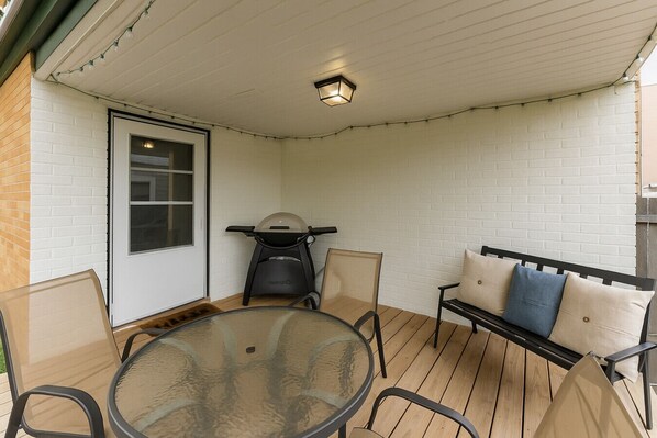 Ein überdachter Patio-Bereich mit einem runden Glastisch, vier Stühlen, einer Bank mit Kissen und einer Deckenleuchte.