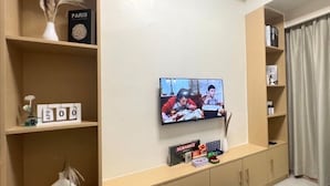 Uma sala de estar com uma TV de tela plana na parede, estantes de madeira, um tapete peludo preto e diversos objetos decorativos, incluindo um vaso com plantas secas e um calendário marcado com ABR 00.