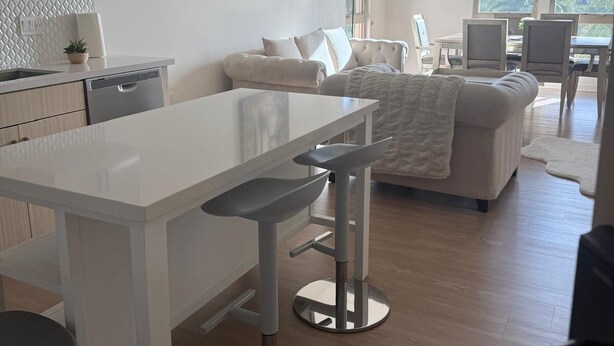 Eine moderne Küche mit weißer Arbeitsplatte, Barhocker und einer Essecke mit einem weißen Sofa und einem Bild an der Wand.