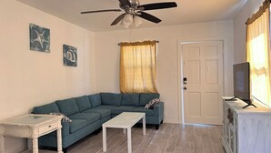 Ein Wohnzimmer mit Ecksofa, einem kleinen weißen Tisch, Deckenventilator, Fernseher auf einem Schrank und zwei Wandbildern.