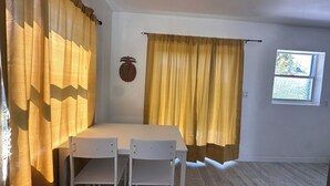 Ein Zimmer mit weißem Tisch und Stühlen, gelben Vorhängen, einer Wanddekoration mit Ananas und einem Fenster mit Blick auf Grün.