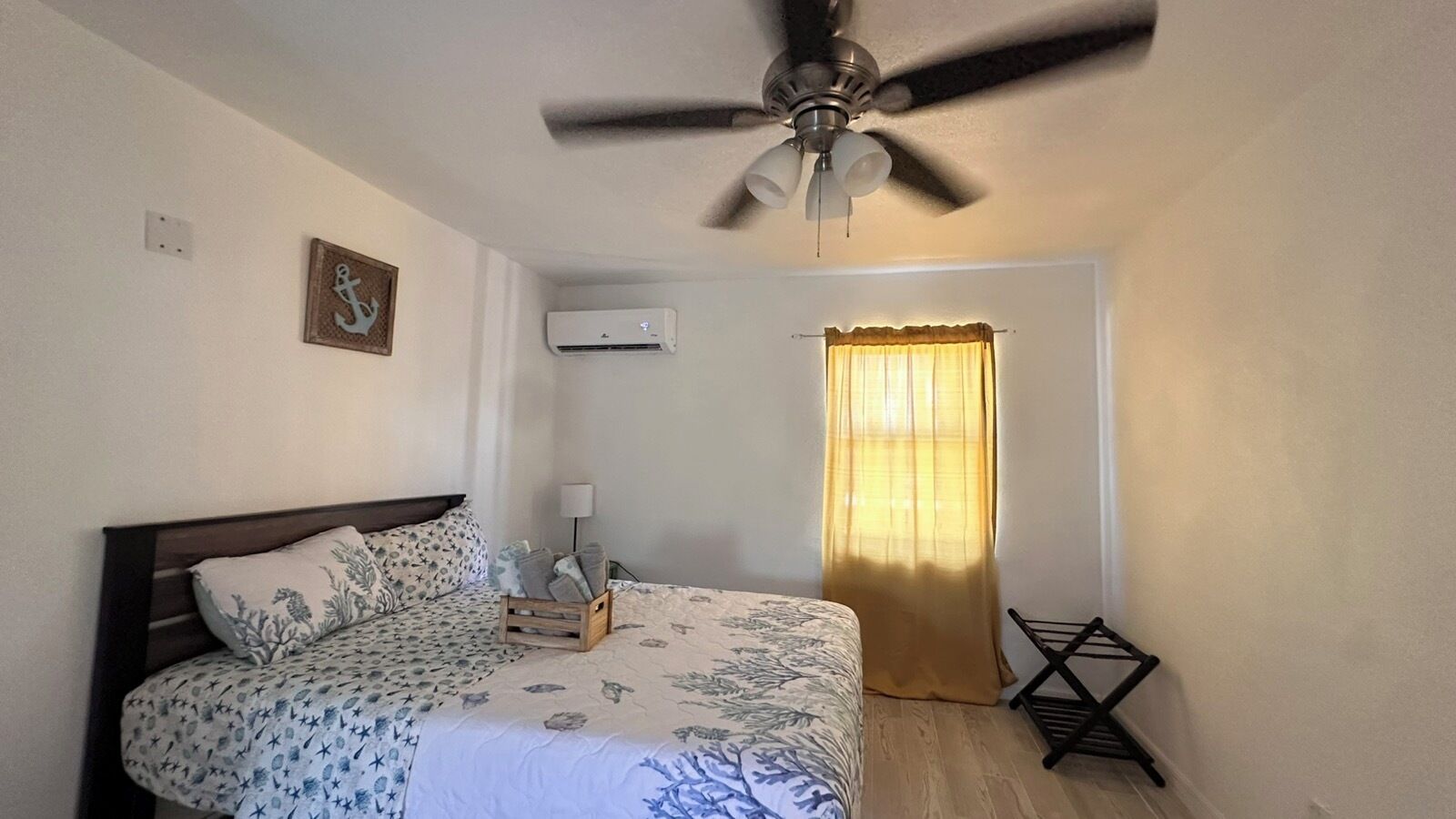 Une chambre à coucher comprenant un lit, un ventilateur de plafond, un climatiseur, une fenêtre avec des rideaux et une petite table avec une lampe.