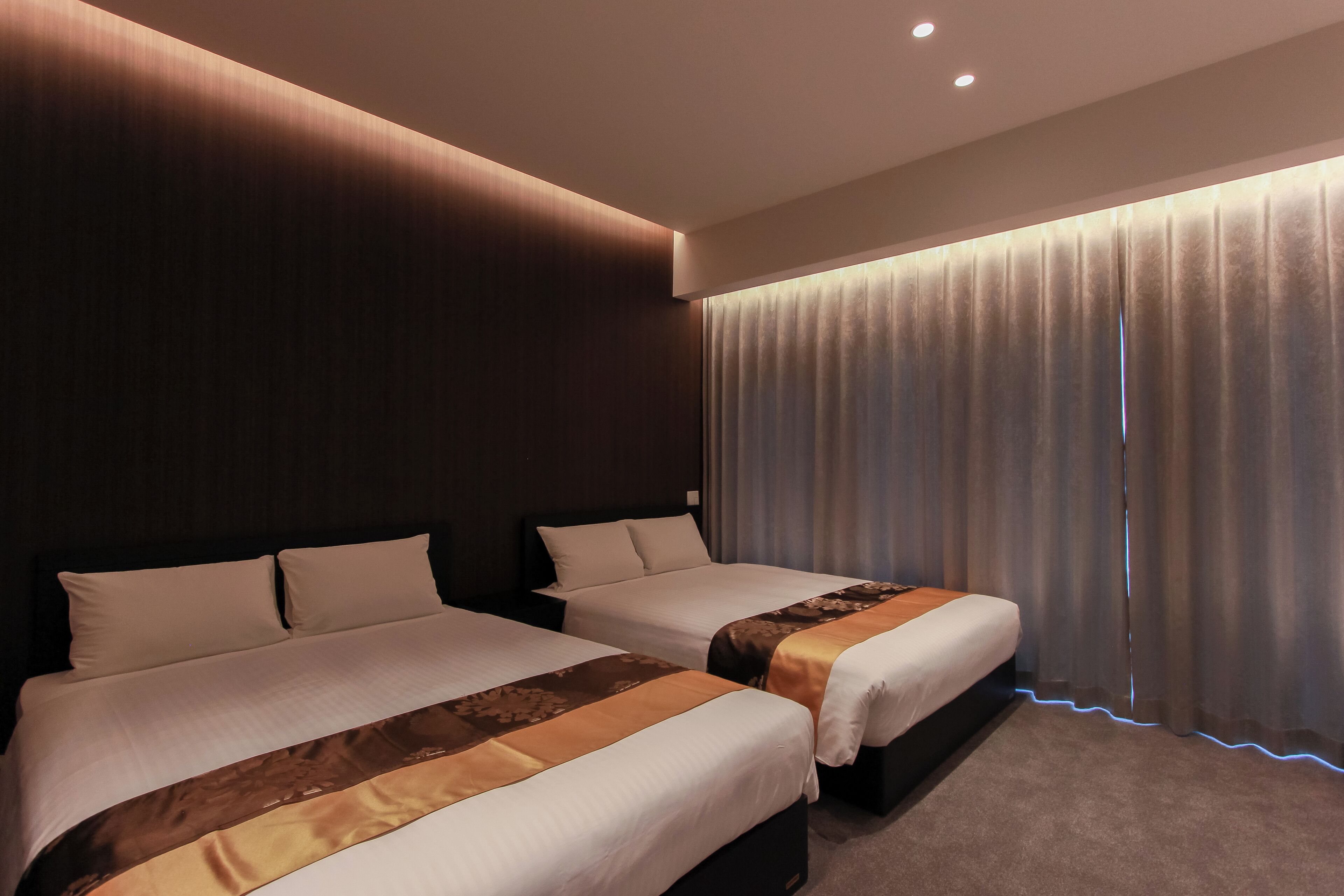 Hotellihuone, jossa on kaksi sänkyä, tumma puinen pääty, suuri ikkuna verhoilla ja matolla peitetty lattia.