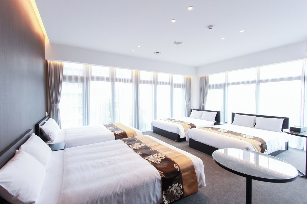 A hotel room with three beds, a small table, and large windows with curtains.
