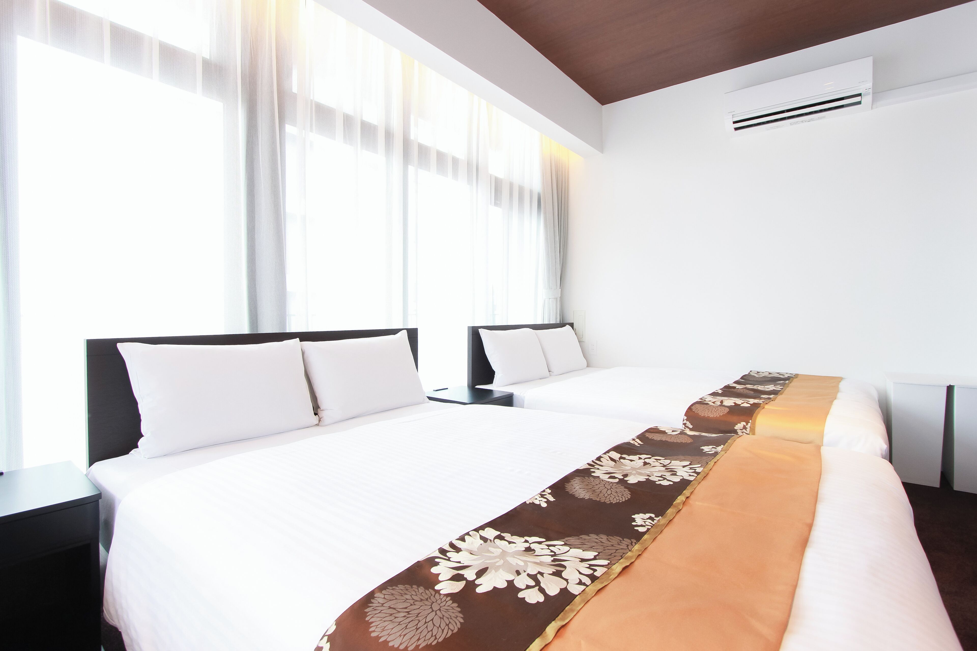 A hotel room with two beds, a large window with sheer curtains, a white air conditioning unit, and a brown decorative runner on the bed.