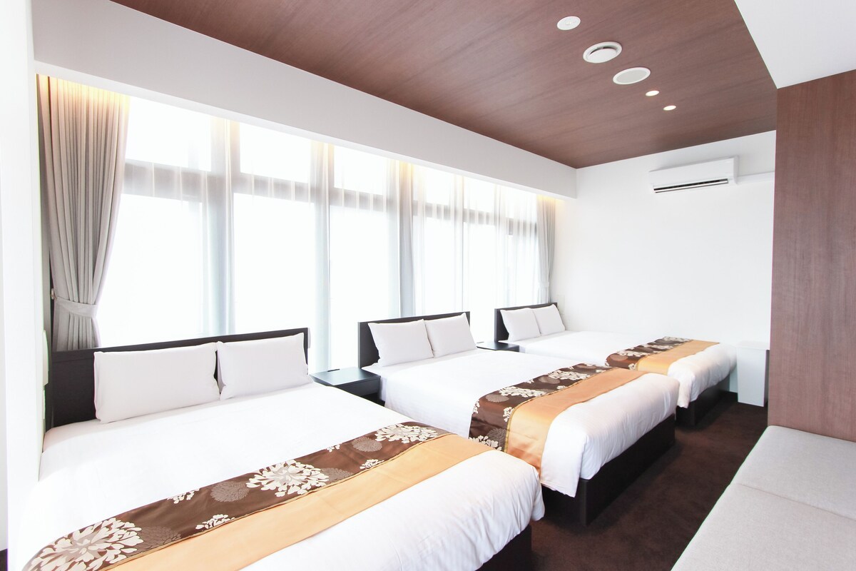 A hotel room with three beds, large windows with sheer curtains, a wooden ceiling, and an air conditioning unit.