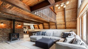Un loft amplio con vigas de madera a la vista, una chimenea, un sofá seccional gris y una cocina al fondo.