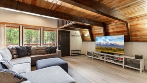 Una sala de estar estilo loft acogedora, con una televisión grande, vigas de madera, un sofá y vistas al exterior.