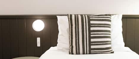 Una cama bien hecha con ropa de cama blanca y una almohada a rayas, una mesita redonda al lado y una lámpara de pared.