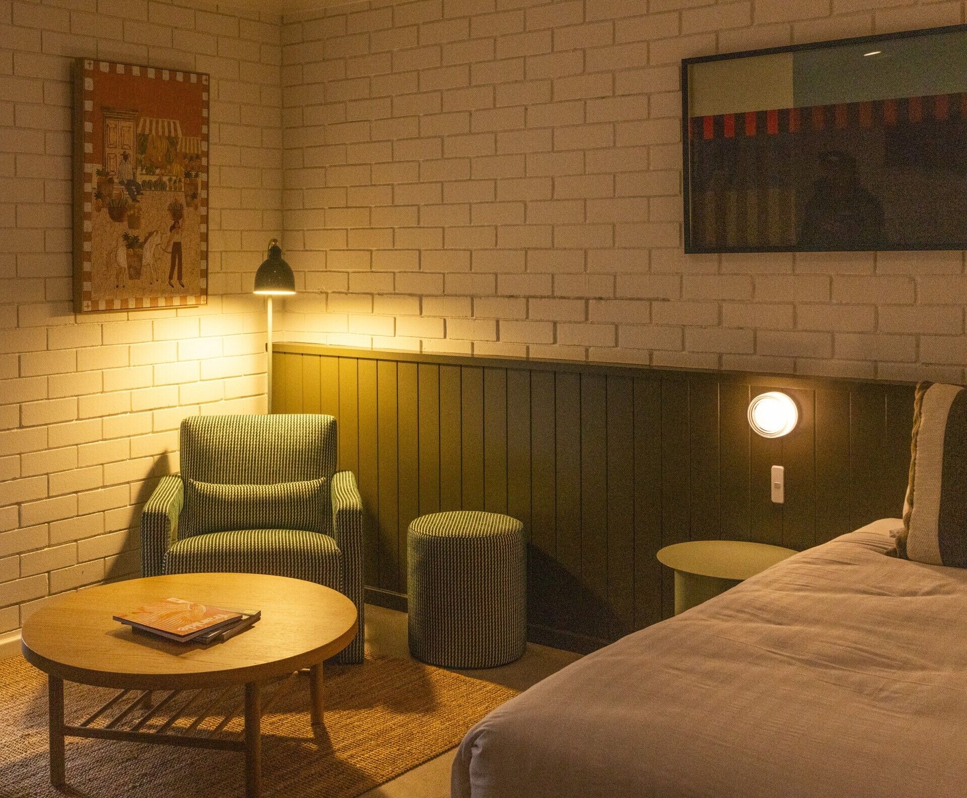 A cozy room with a brick wall, a green-striped armchair, a round wooden table, a small stool, and a bed with a headboard.
