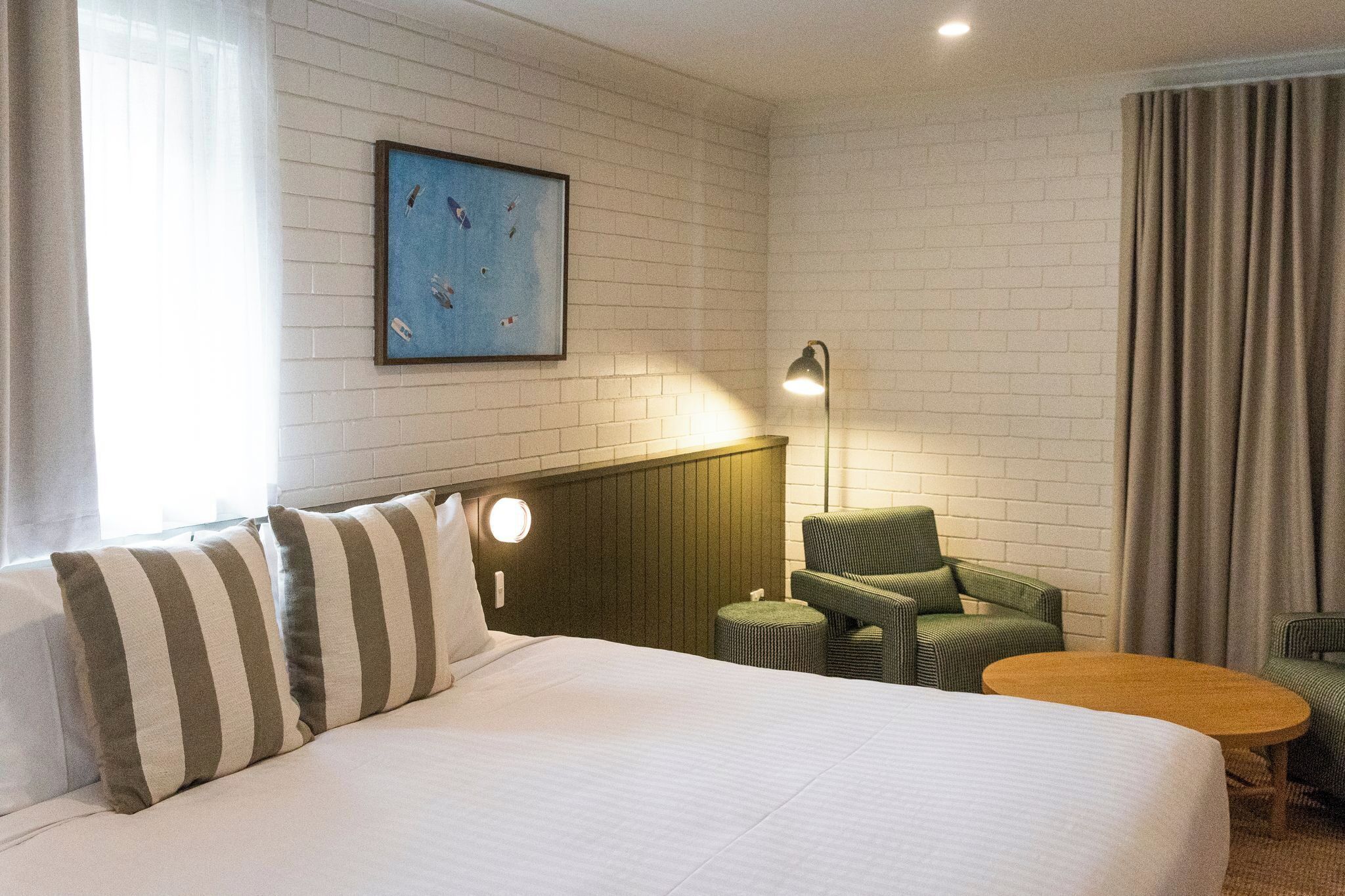 A hotel room with a large bed, a round table, two armchairs, a small stool, a wall-mounted lamp, and a framed picture on the wall.