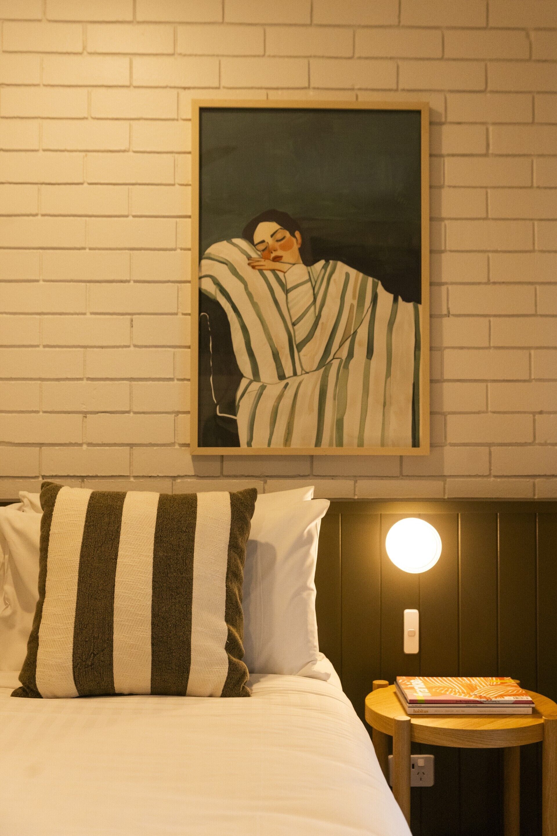 Ein ordentlich bezogenes Bett mit gestreiften Kissen, ein gerahmtes Kunstwerk an der Wand und ein Nachttisch mit einem Buch und einer Lampe.