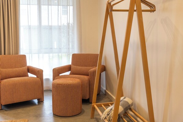Una habitación con dos sillones marrones de mimbre, un pequeño otoman y una mecedora de madera, además de una ventana con cortinas de gasa.