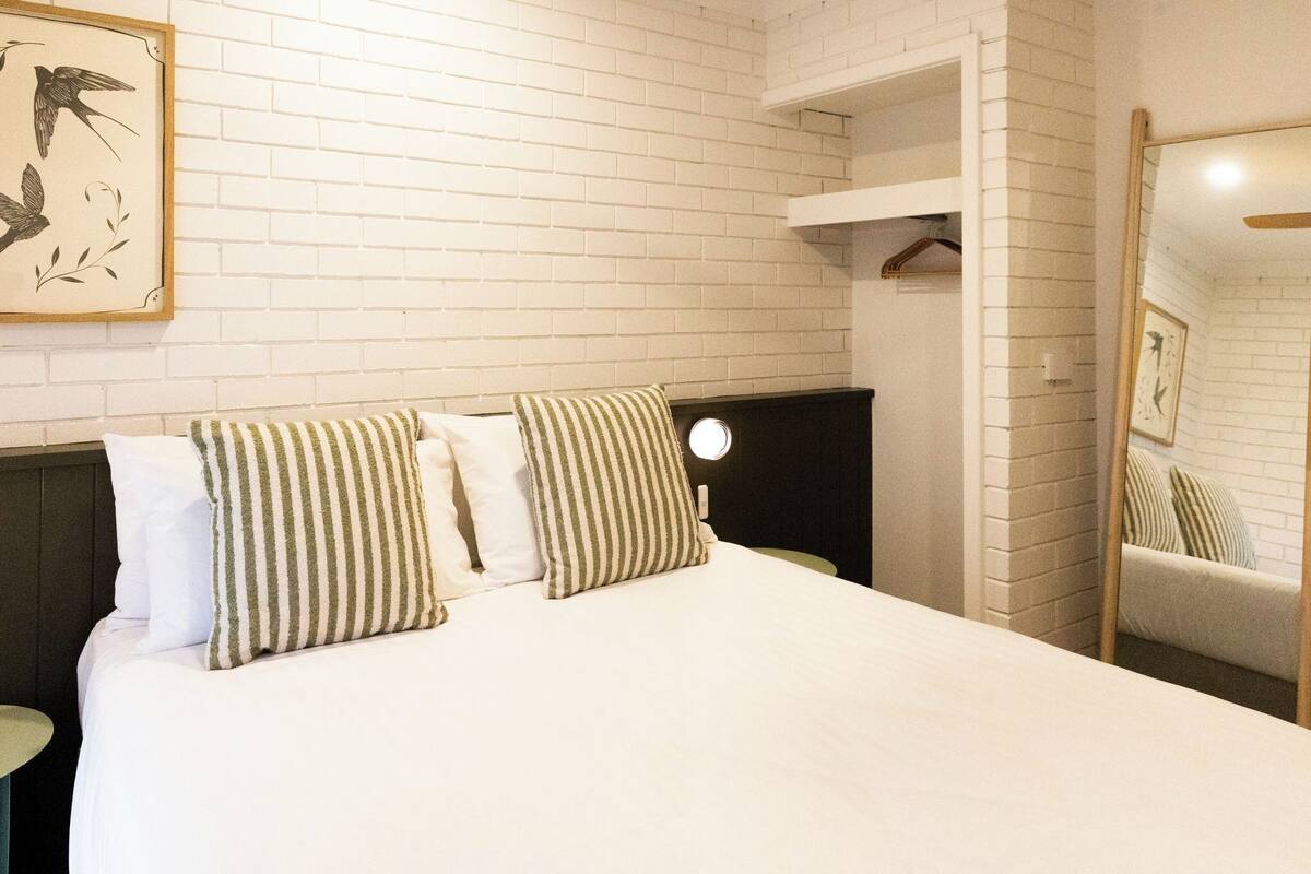 A hotel room with a white brick wall, a bed with striped pillows, a round night light, and a mirror reflecting the room.