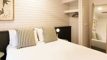 Een hotelkamer met een witte bakstenen muur, een bed met gestreepte kussens, een ronde nachtlamp en een spiegel die de kamer weerspiegelt.