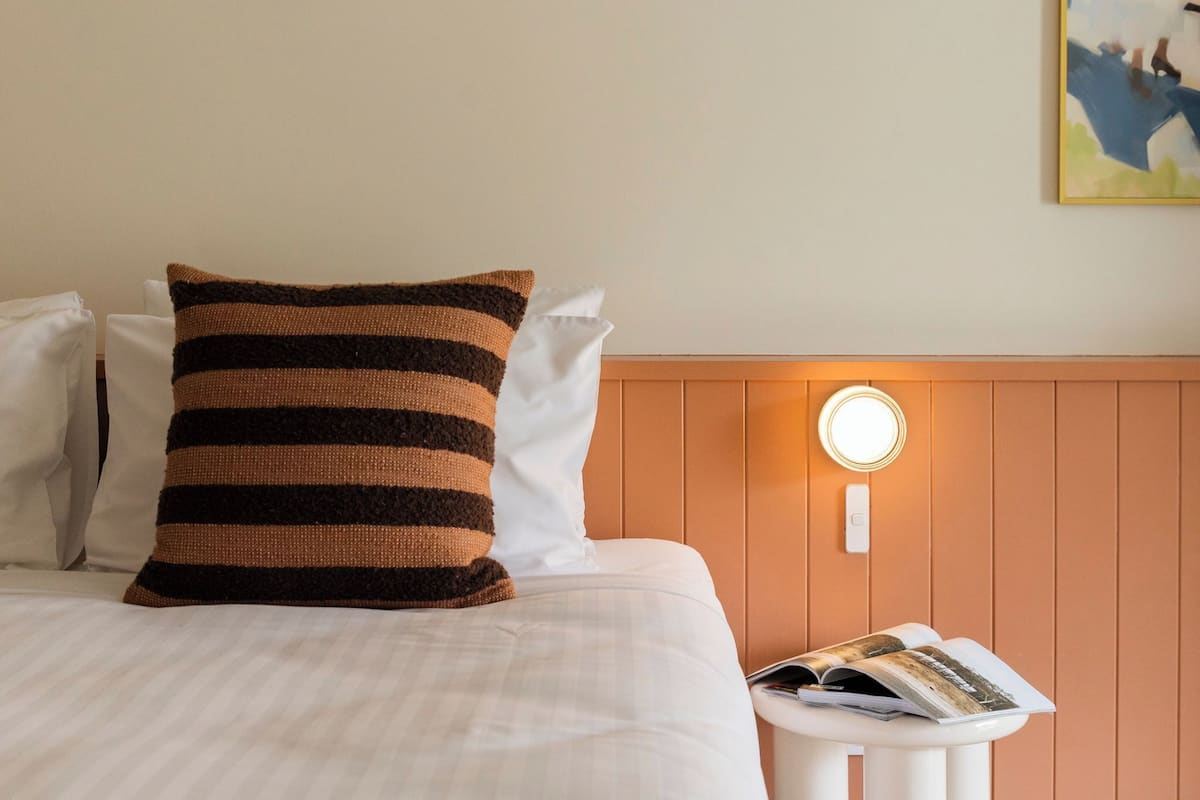 A neatly made bed with striped pillows, a bedside table with a magazine, and a wall-mounted light.