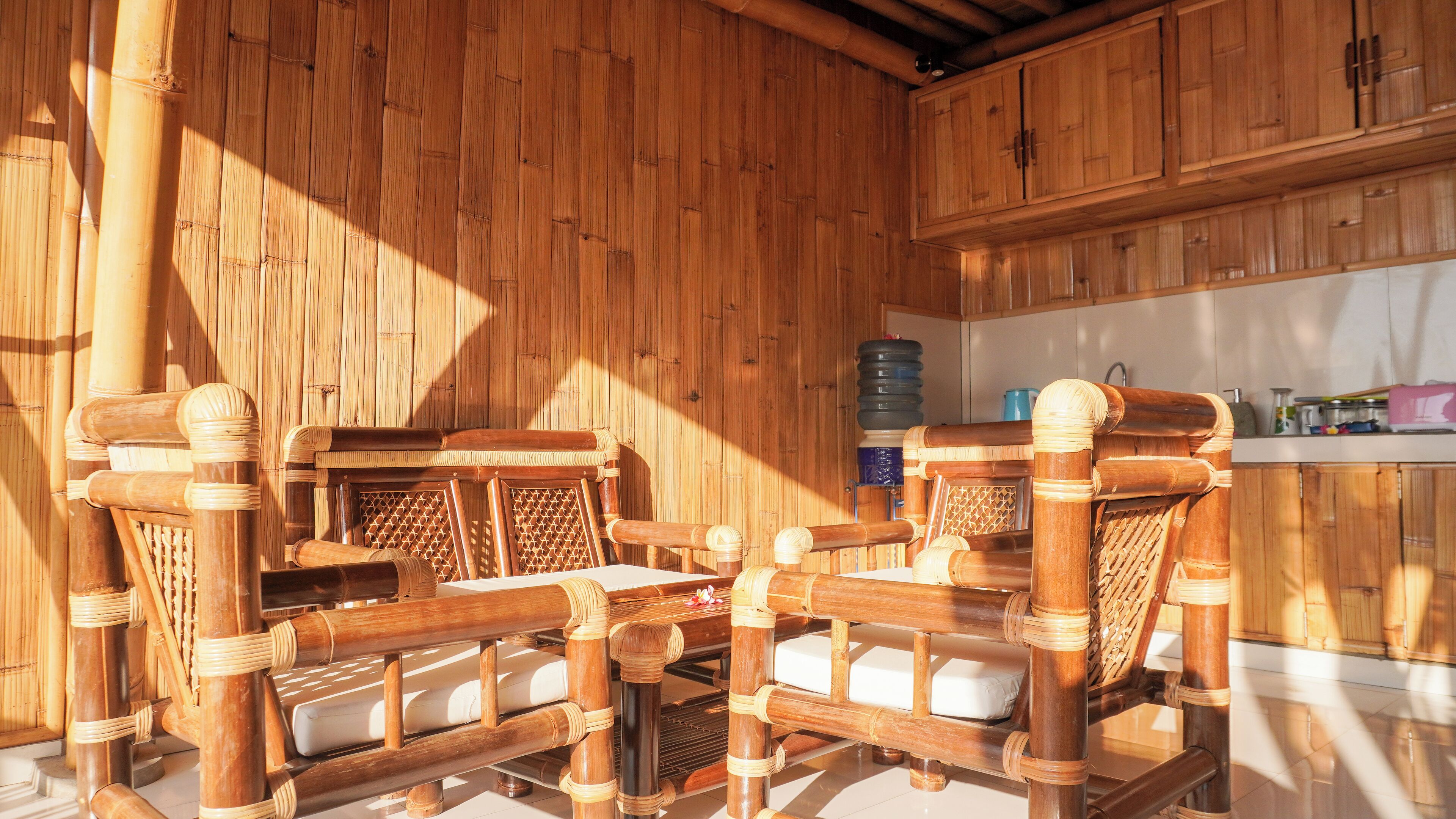 Un área de estar con estructura de bambú, con sillones acolchados y una mesa, una cocina con armarios y encimera, y un dispensador de agua.