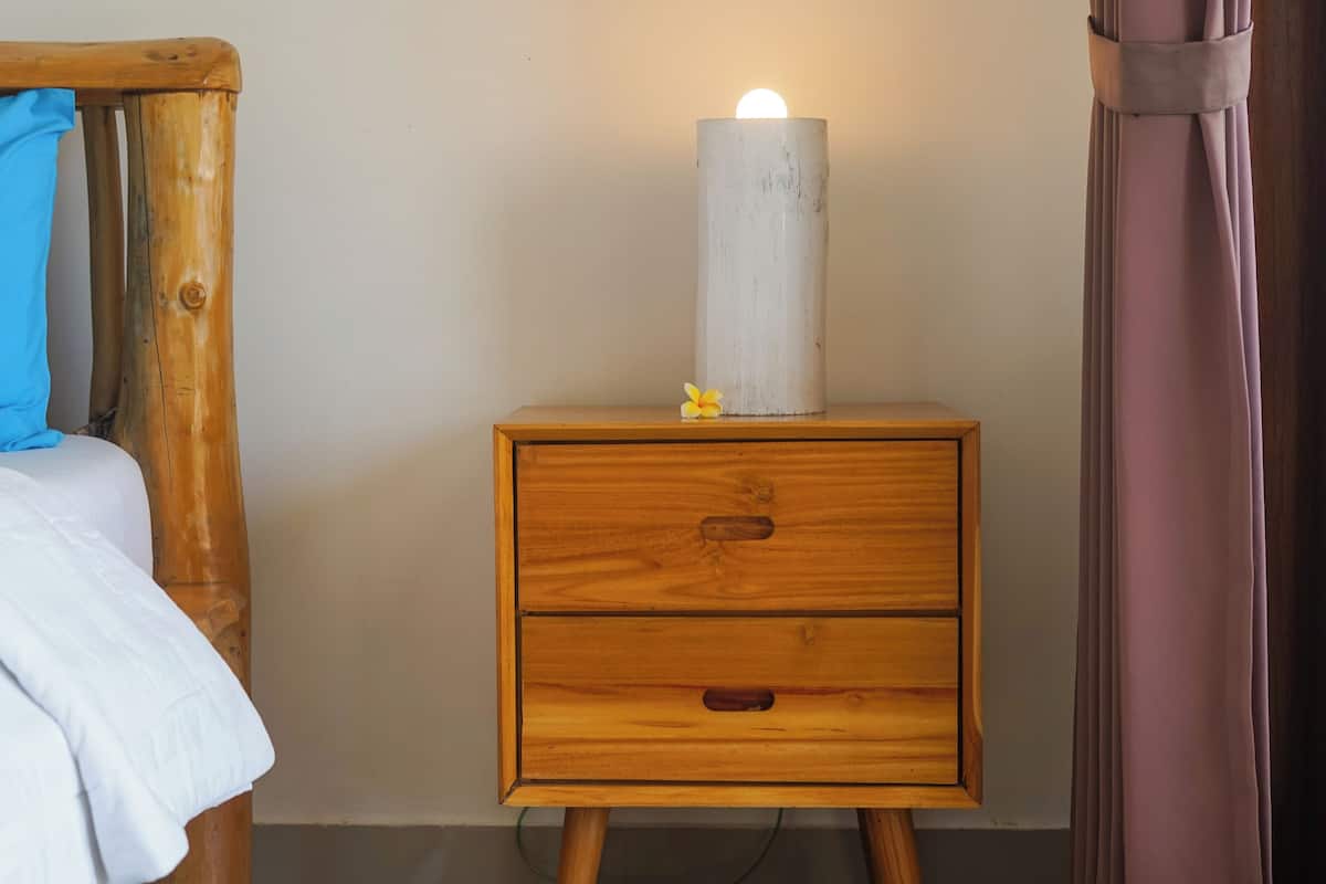 Ein Nachttisch aus Holz mit einer weißen Kerze und einer kleinen Blume, ein Bett aus Holz mit einem blauen Kopfkissen und ein Nachttisch aus Holz mit einer Schublade.
