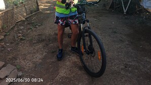Une personne se tient à côté d’un vélo dans un paysage rural, avec une maison et des arbres en arrière-plan.