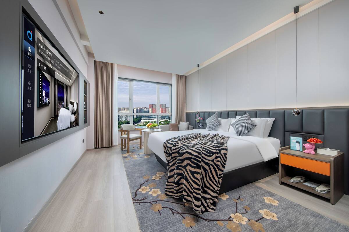 A modern hotel room with a large bed, a bedside table, a TV, and a city view.