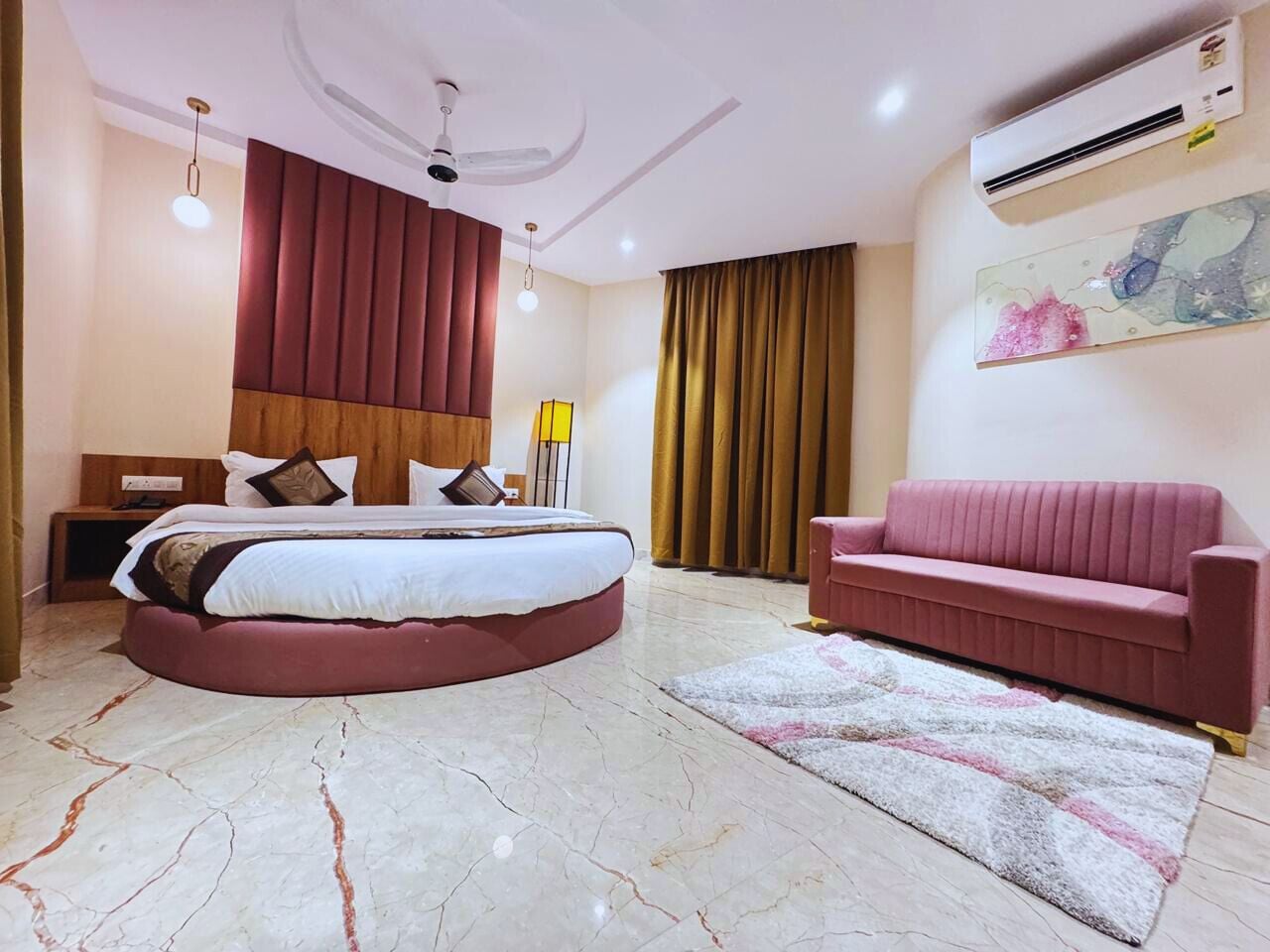 A bedroom with a round bed, a pink sofa, a ceiling fan, an air conditioner, and a large window with curtains.