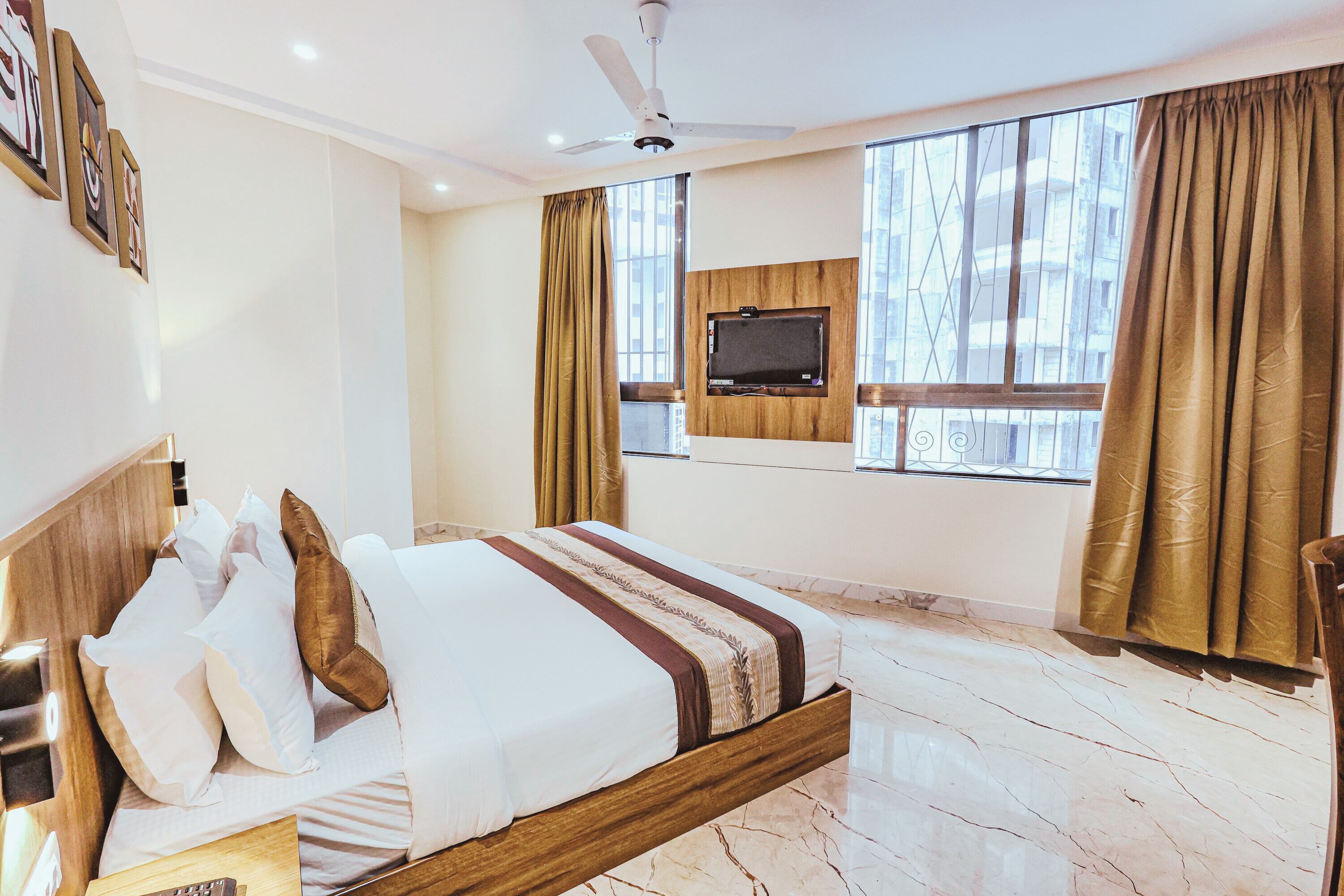 A hotel room with a bed, a ceiling fan, a TV mounted on the wall, and large windows with curtains.