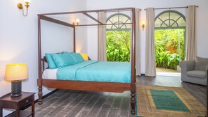 Una cama con dosel, con un edredón y almohadas color turquesa, una mesita de noche de madera con una lámpara, un sofá gris y vistas a zonas verdes a través de una ventana con cortinas.