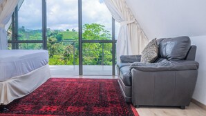 Una habitación con una cama, un sofá gris, un tapete rojo estampado y un ventanal con vistas a zonas verdes.
