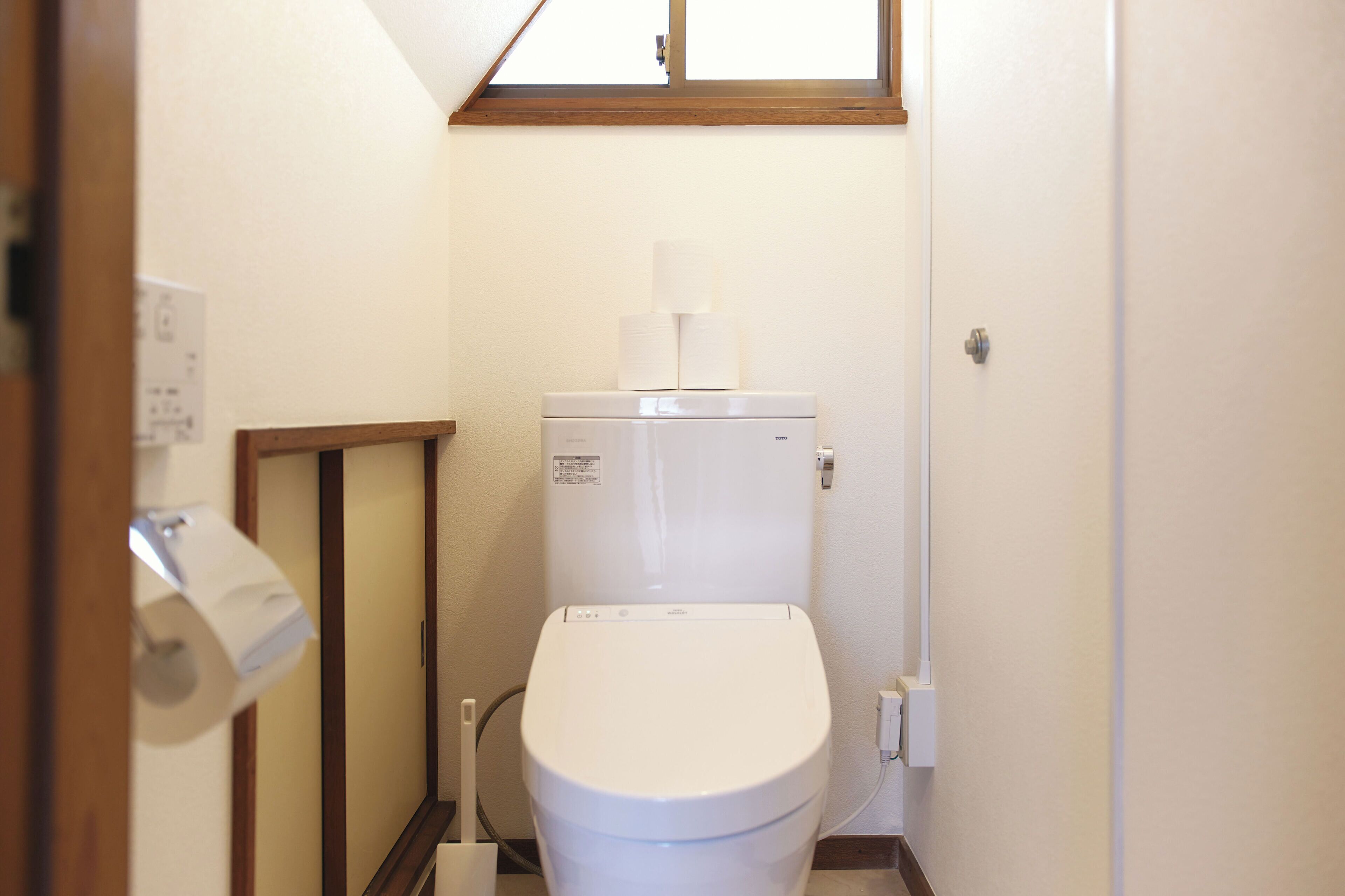 A small, clean bathroom with a toilet, a roll of toilet paper, and a window above the toilet.