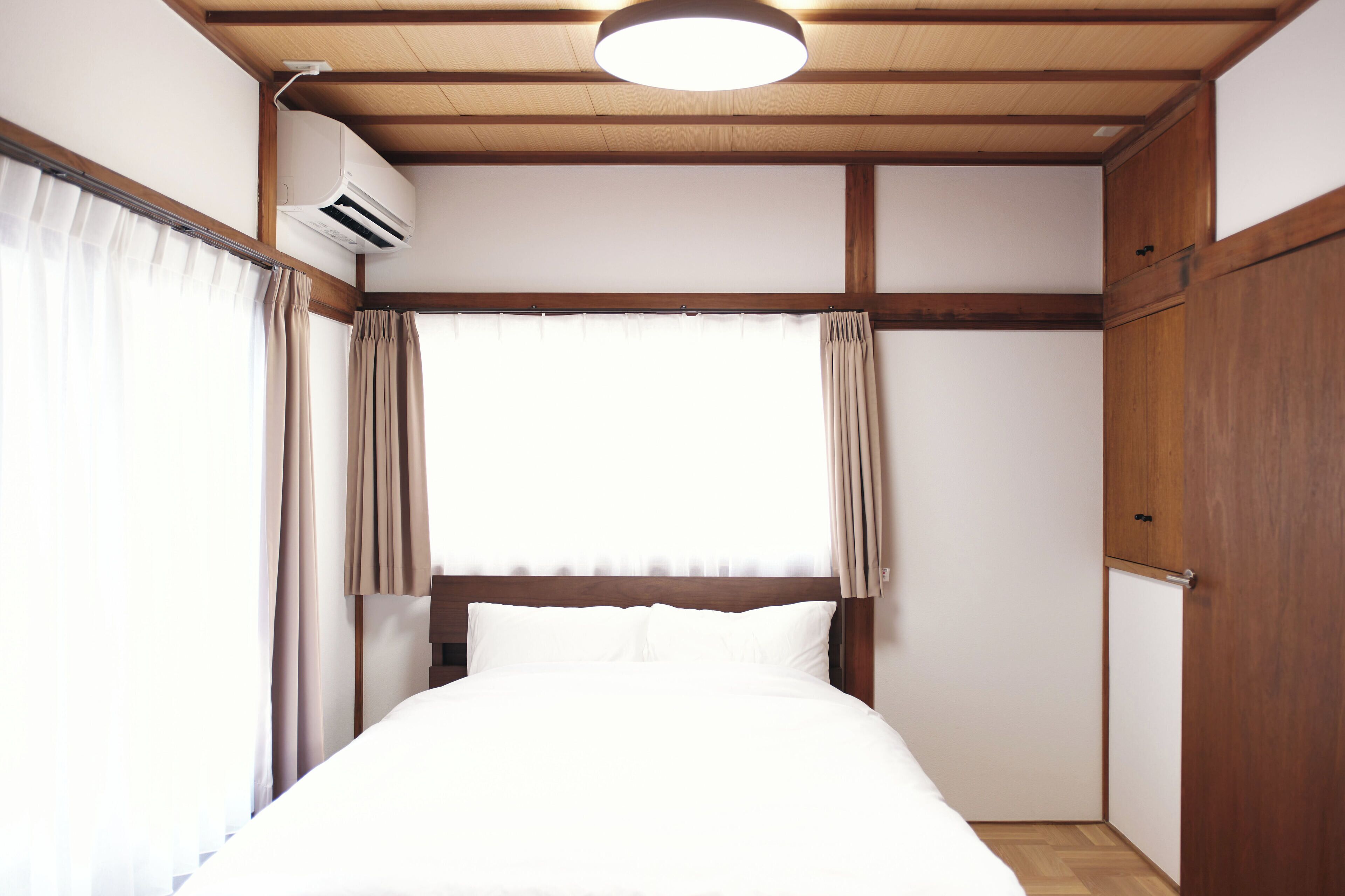 A hotel room with a bed, wooden ceiling beams, an air conditioning unit, and a window with curtains.