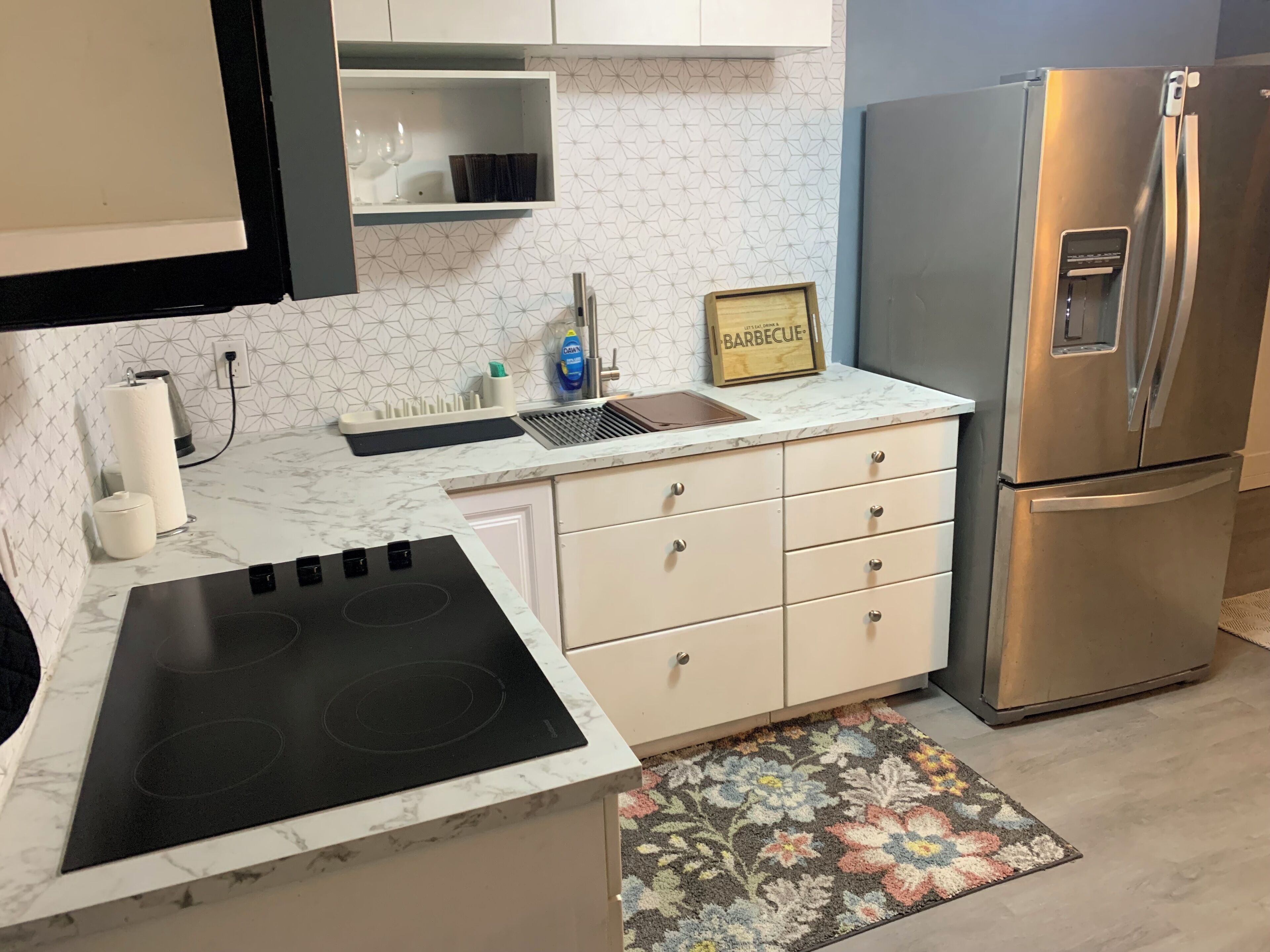 A kitchen with a stainless steel refrigerator, a black induction cooktop, white cabinets, and a marble countertop.