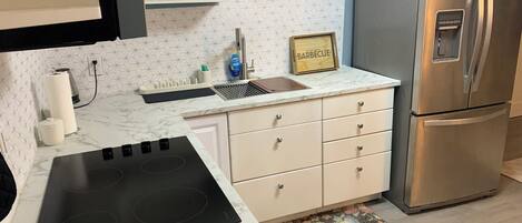 A kitchen with a stainless steel refrigerator, a black induction cooktop, white cabinets, and a marble countertop.
