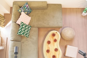 Una habitación con un sofá verde, cojines estampados, un pouf tejido, un cabecero de madera y un taburete de madera pequeño.