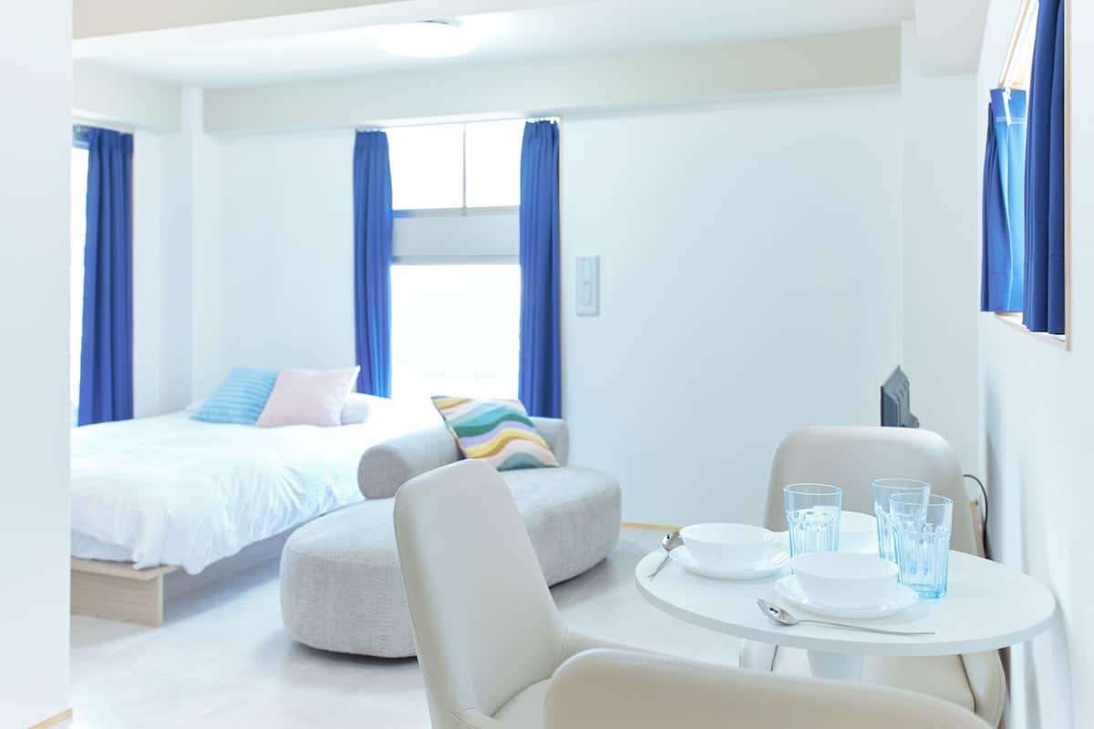 A modern hotel room with a bed, a sofa, a dining table with glasses and plates, and a window with curtains.